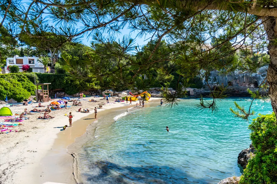 Sandbucht in Cala d'Or zwischen hellen Felswänden, davor sehr klares türkisfarbenes Wasser mit Badegästen.