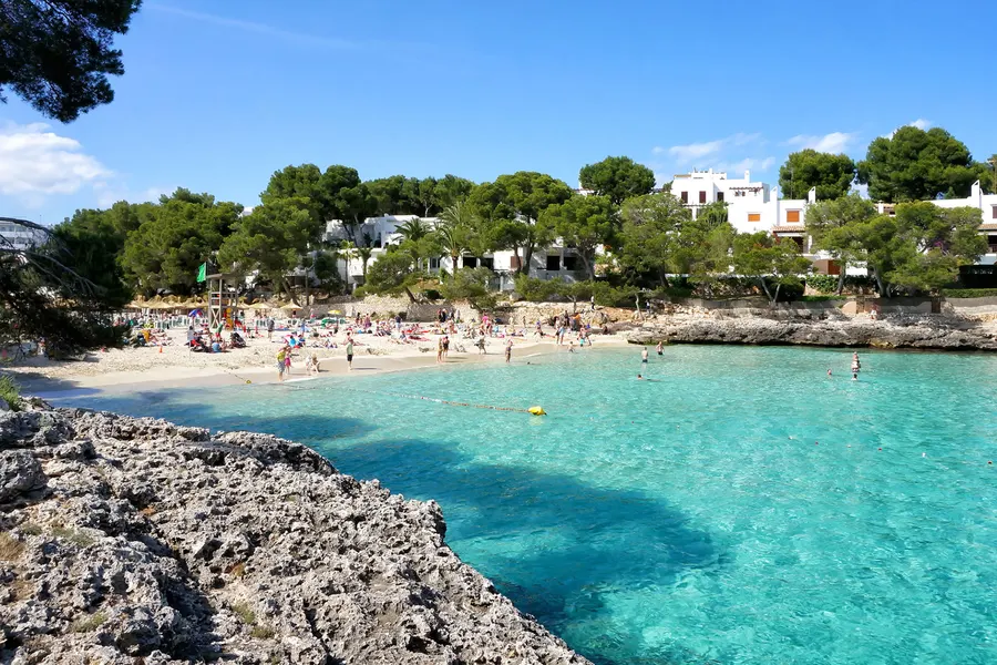Blick von der Felskante über eine Bucht von Cala d'Or mit Badegästen im flachen Wasser, dahinter weiße Ferienhäuser zwischen Kiefern.