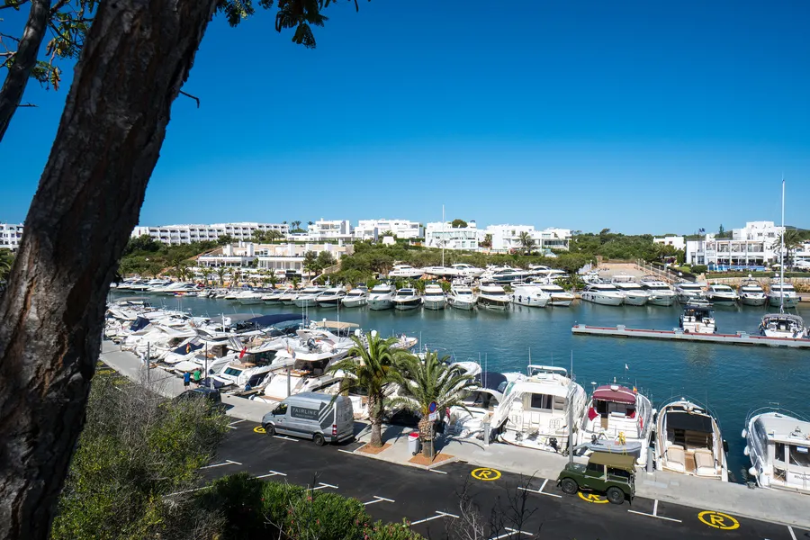 Yachthafen von Cala d'Or mit dicht liegenden Booten, Palmen am Kai und der weißen Bebauung am gegenüberliegenden Ufer.

