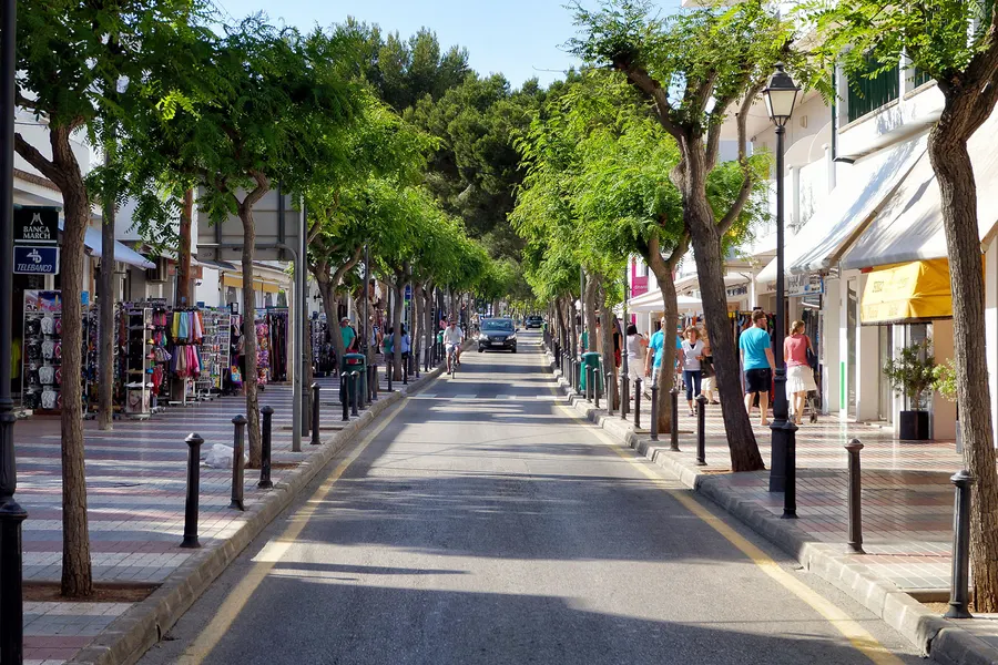 Einkaufsstraße in Cala d'Or mit Läden, Markisen, Bäumen am Straßenrand und Passanten unter blauem Himmel.
