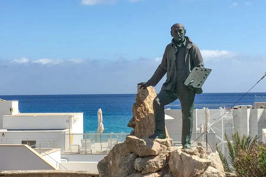 Bronzestandbild eines Malers mit Palette in der Hand auf einem Felssockel über dem Meer, dahinter das offene Wasser und weiße Terrassen.
