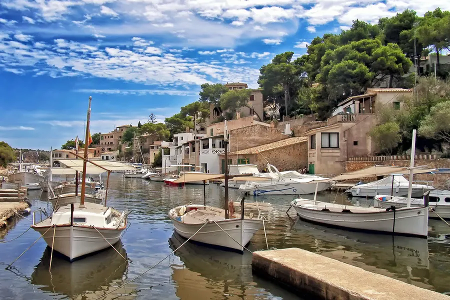 Weiße Llaüts, traditionelle mallorquinische Fischerboote, im ruhigen Wasser des Hafenarms von Cala Figuera, dahinter Bootshäuser und Kiefern.