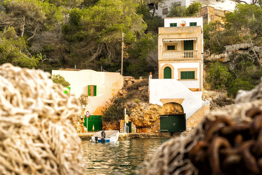 Blick über unscharfes Tauwerk hinweg auf ein steinernes Bootshaus mit grünen Fensterläden am Wasser von Cala Figuera.
