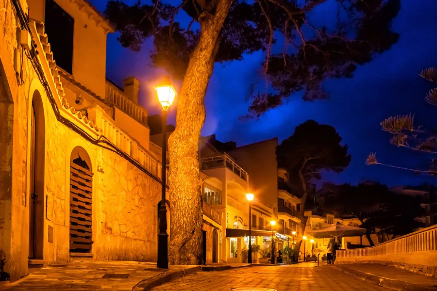 Beleuchtete Gasse in Cala Figuera bei Nacht mit Straßenlaterne, einer alten Kiefer und Sandsteinmauern, dahinter Lichter am Wasser.
