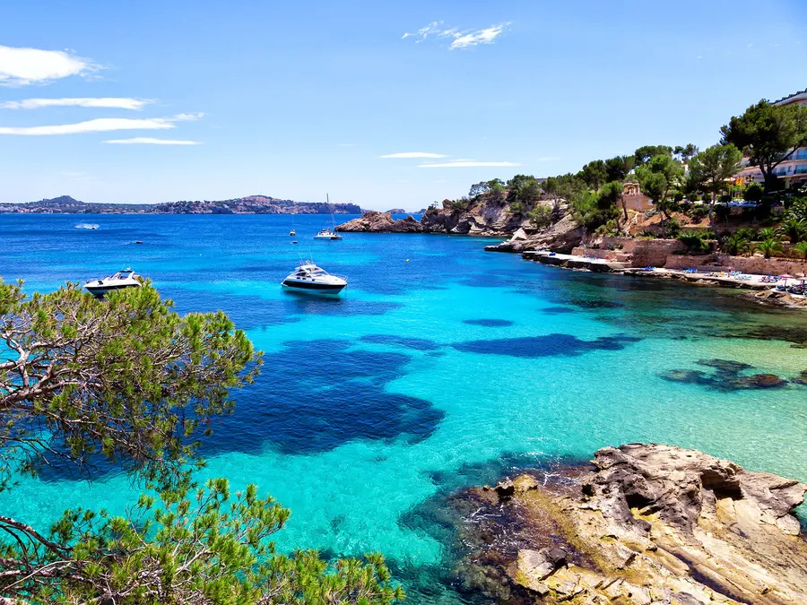 Flaches Felsufer der Cala Fornells mit sehr klarem, türkis- und blaugrün schimmerndem Wasser, im Hintergrund eine bewaldete Landzunge.
