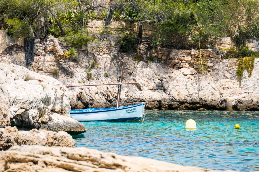 Ein blau-weißes Fischerboot im flachen klaren Wasser einer kleinen Felsbucht bei Cala Fornells, ringsum Kiefern und heller Fels.