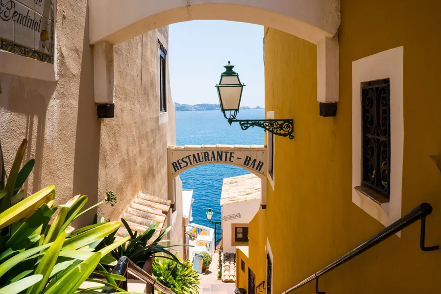 Schmale Gasse zwischen ockerfarbenen Häusern in Cala Fornells mit schmiedeeiserner Laterne und einem Restaurantschild, am Ende der Blick aufs Meer.
