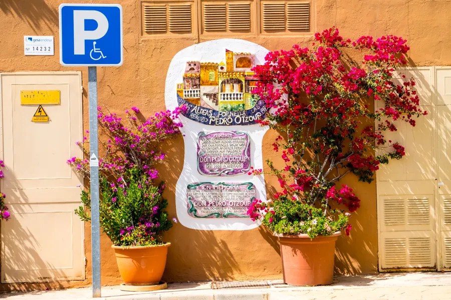 Leuchtend pinke Bougainvillea an einer sandfarbenen Hauswand in Cala Fornells, davor Blumentöpfe und ein Hinweisschild.
