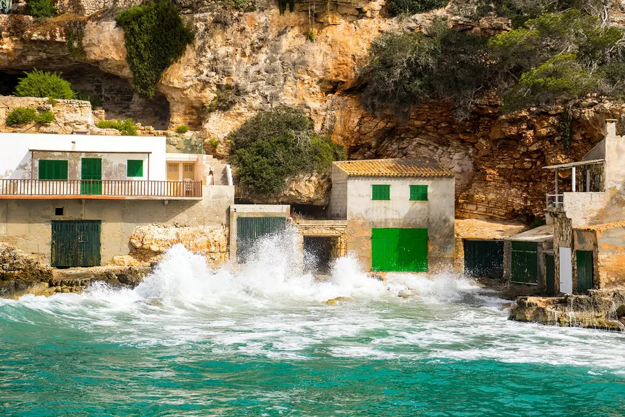 Hoch aufspritzende Gischt vor den in den Fels gebauten Bootshäusern der Cala Llombards bei kräftigem Wellengang.
