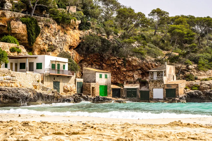 Blick von oben auf die Reihe der Fischerhäuser am Felsrand der Cala Llombards, davor auslaufende Wellen und heller Fels.

