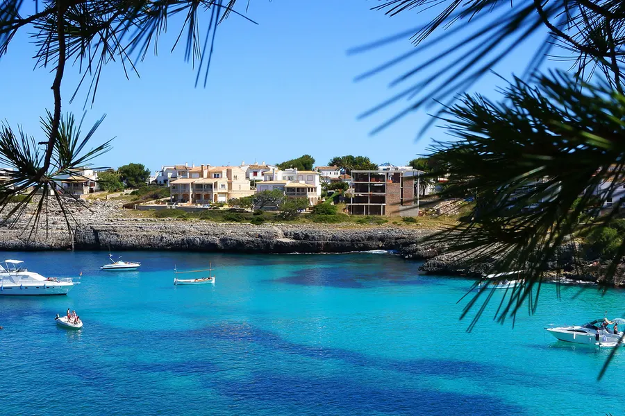 Blick durch Kiefernzweige auf die Bucht der Cala d'en Marçal mit ankernden Motorbooten, tiefblauem Wasser und den Apartmentbauten am Ufer.
