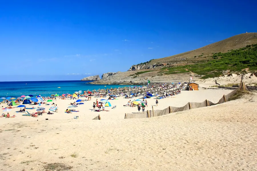 Dicht belegter Strand der Cala Mesquida mit bunten Sonnenschirmen und Badegästen, dahinter der bewachsene Hang und blauer Himmel.
