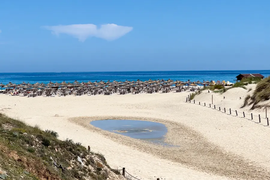 Reihe von Schilfsonnenschirmen am Rand des Strands, davor eine flache wassergefüllte Senke im Sand, dahinter die Dünen.
