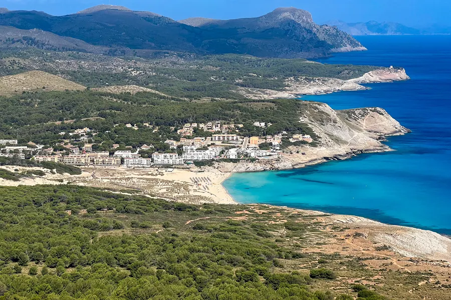 Blick über die bewaldeten Hügel auf die Siedlung von Cala Mesquida und die türkisfarbene Bucht, im Hintergrund die Steilküste und das offene Meer.

