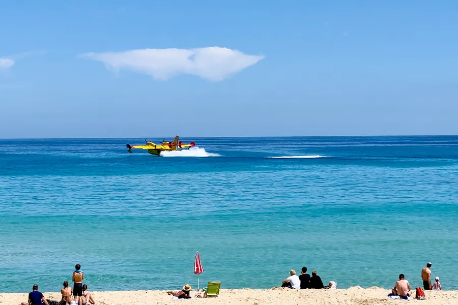 Löschflugzeug dicht über der Wasseroberfläche vor der Cala Mesquida beim Aufnehmen von Wasser, im Vordergrund Badegäste am Ufer.
