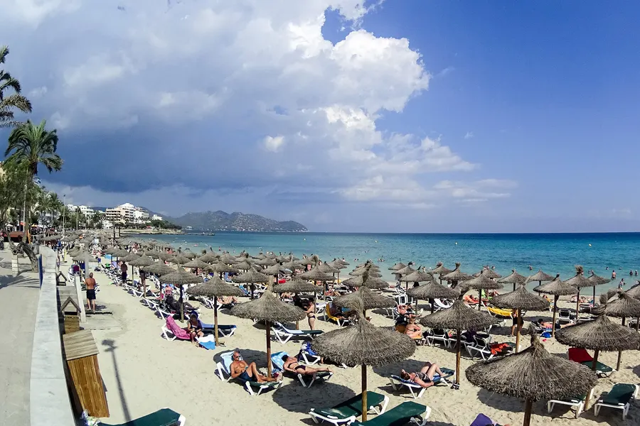 Blick über die Promenade von Cala Millor mit Strohschirmen am Strand, Palmen und Hotelbauten, dahinter die Berge des Ostens.