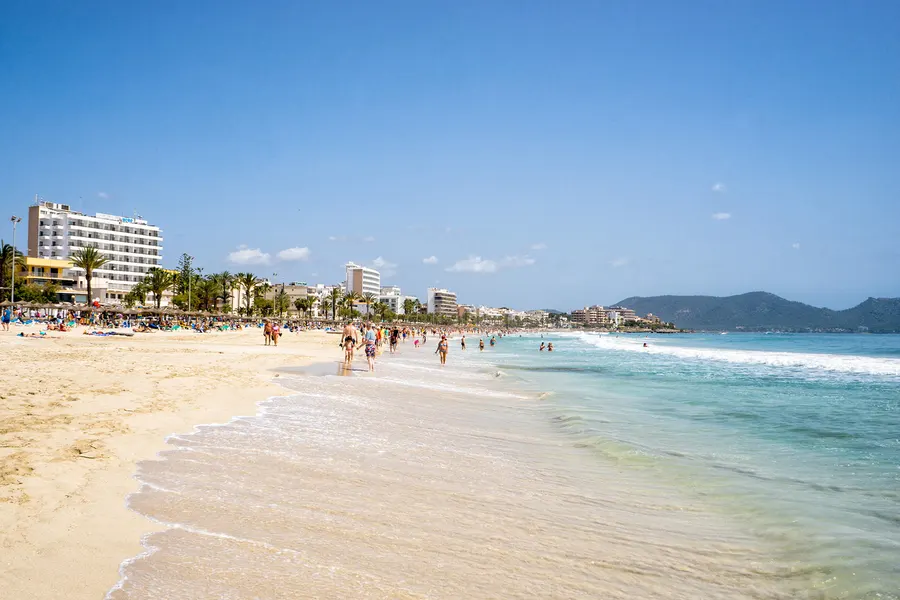 Sehr klares türkisfarbenes Flachwasser vor Cala Millor mit sandigem Grund, dahinter der Strand und die Häuserzeile.
