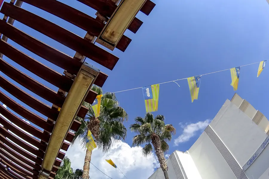 Besetzte Terrasse eines Lokals in Cala Millor unter großen Bäumen mit Sonnenschirmen und roten Stühlen.
