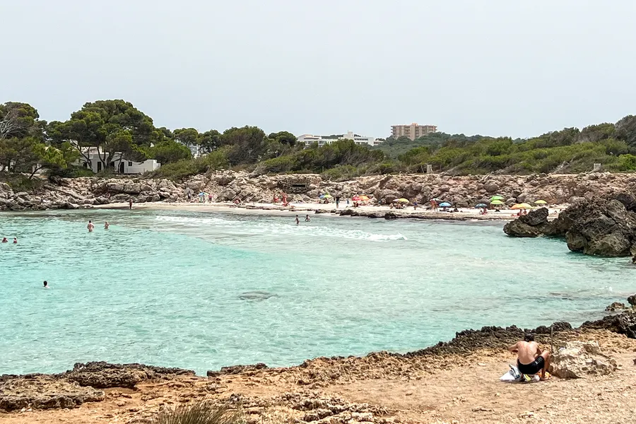 Blick über die zerklüftete Felsküste bei der Cala Moltó auf türkisfarbenes Wasser und die bewaldeten Hänge dahinter.