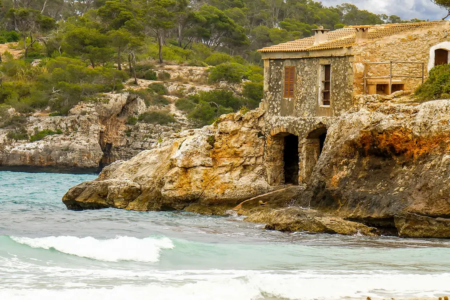 Altes zweistöckiges Steinhaus auf dem Fels über einer Bucht der Cala Mondragó, davor auslaufende Wellen und heller Sand.
