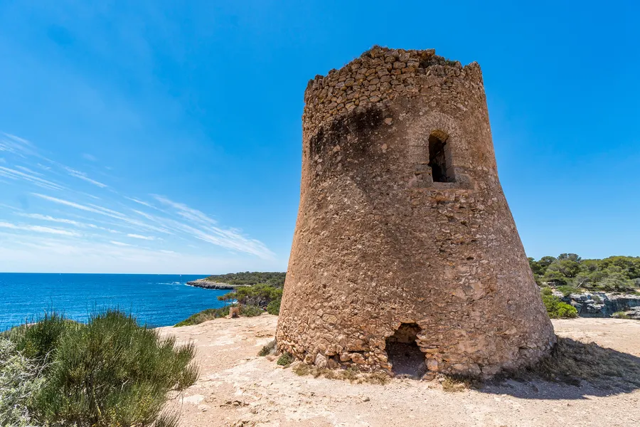 Der runde Wachturm Torre de Cala Pi aus grobem Bruchstein auf der Klippe, dahinter das offene Meer unter blauem Himmel.