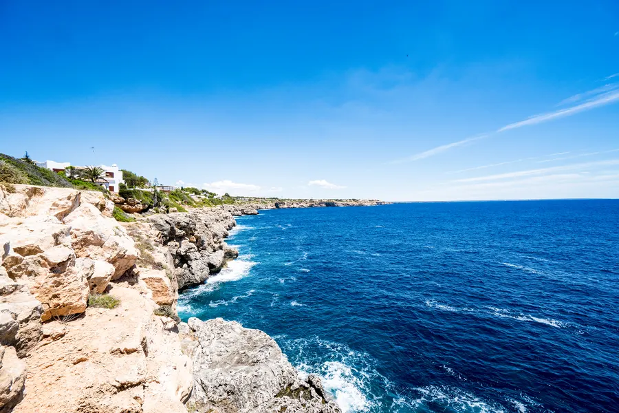 Zerklüftete helle Felsküste bei Cala Pi mit tiefblauem Meer und Brandung, im Hintergrund Häuser am Klippenrand.
