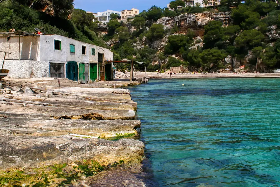 Flache Felsplatten am Ufer der Bucht neben einem alten Bootshaus mit grünen Toren, dahinter das klare grüne Wasser der Schlucht.
