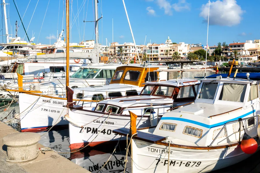 Reihe alter hölzerner Fischerboote mit farbigen Aufbauten und Kennzeichen im Hafen von Cala Ratjada, dahinter die weißen Häuser des Orts.