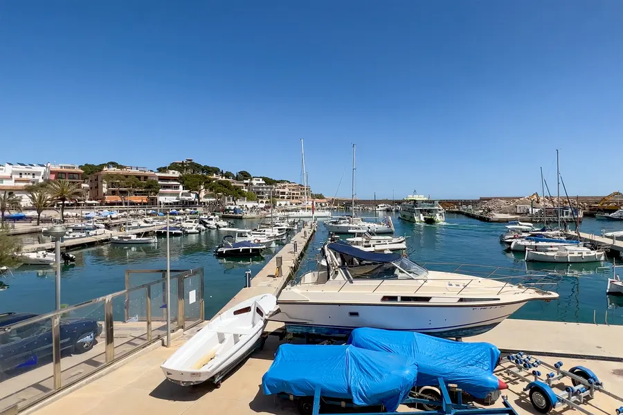 Blick über das Hafenbecken von Cala Ratjada mit Motor- und Segelbooten am Steg, im Hintergrund die bewaldete Landzunge und Häuser am Hang.
