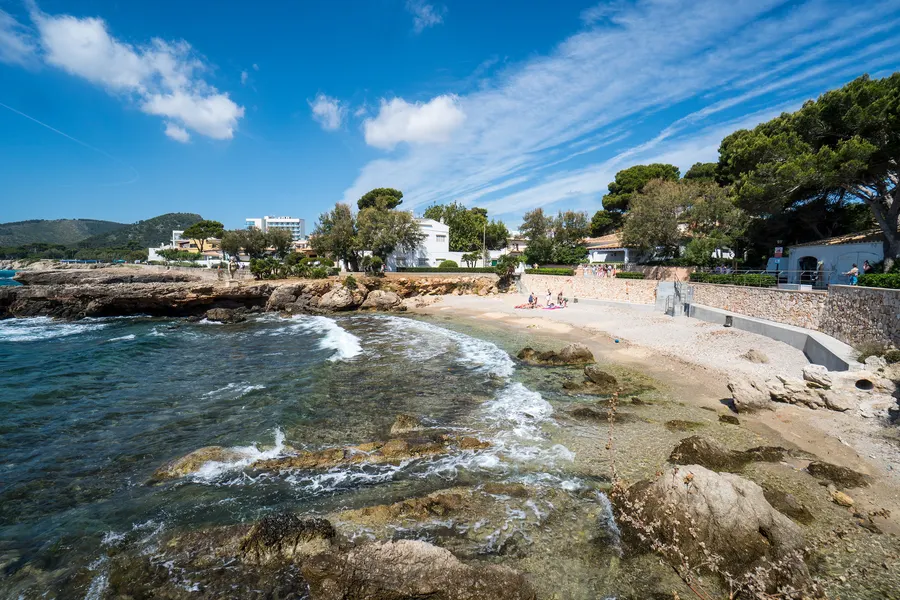 Die Platja de Na Ferradura in Cala Ratjada: kleine Sandbucht mit auslaufender Brandung und Felsen im Vordergrund, dahinter die Hotel- und Häuserzeile.