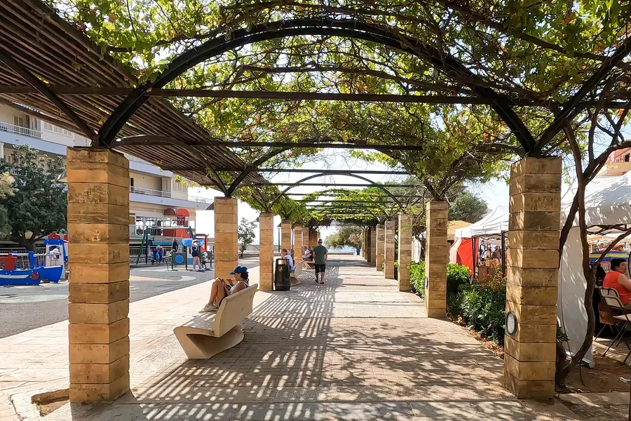 Lange Pergola aus Steinpfeilern und Holzbalken über einem gepflasterten Promenadenabschnitt in Cala Ratjada, mit Bänken im Schatten.
