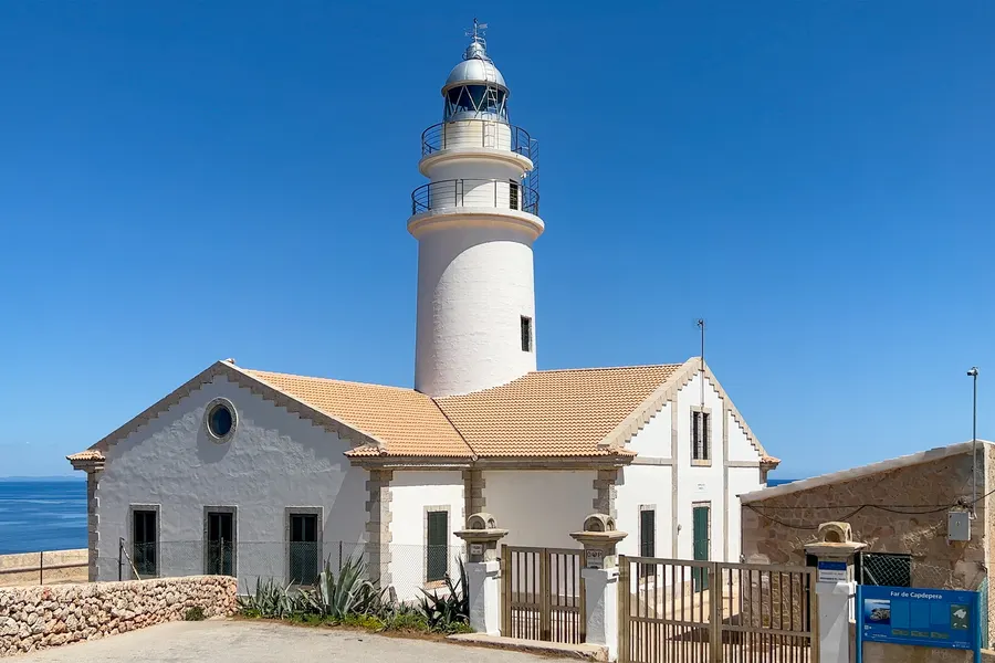 Der weiße Leuchtturm Far de Capdepera mit dem angeschlossenen Wärterhaus hinter einem schmiedeeisernen Zaun, dahinter das offene Meer.
