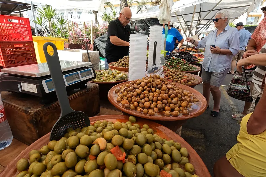 Große Tonschalen mit grünen und eingelegten Oliven auf einem Marktstand in Cala Ratjada, daneben eine Waage, dahinter Kundinnen und Händler unter Planen.
