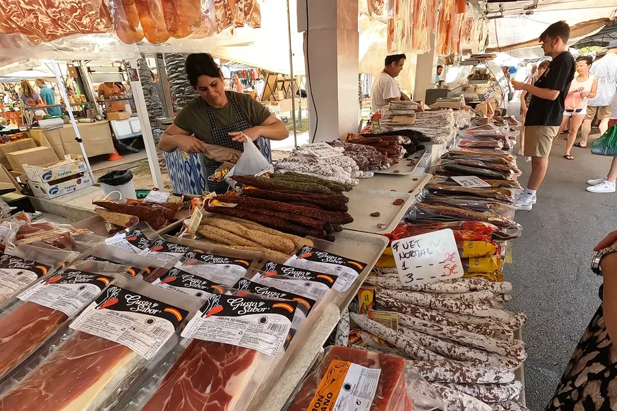Marktstand mit abgepacktem Schinken, hängenden und ausgelegten Würsten, dahinter eine Händlerin beim Einpacken und die Gasse mit weiteren Ständen.
