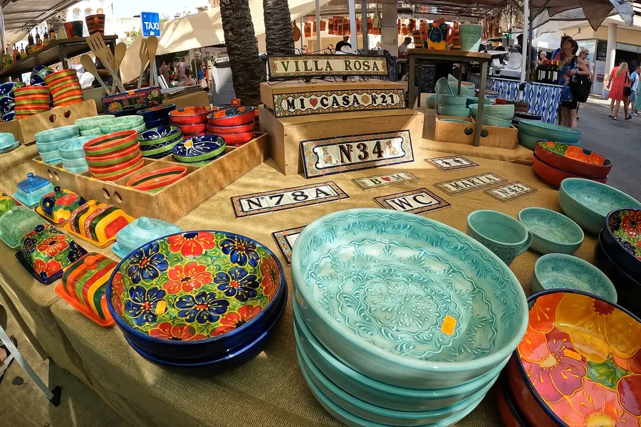 Stand mit bunt bemalter mallorquinischer Keramik, gestapelten türkisfarbenen Schalen und beschrifteten Hausnummernschildern auf dem Markt in Cala Ratjada.
