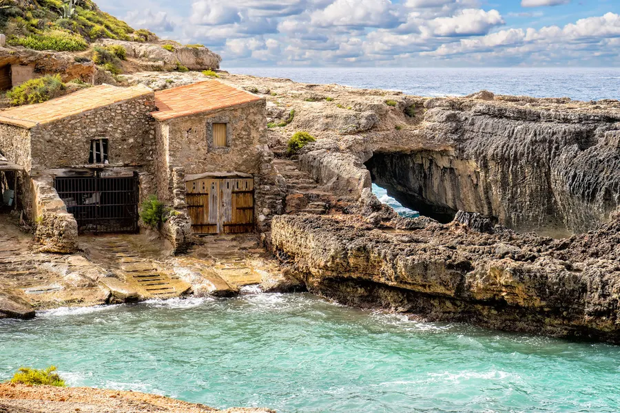 Altes steinernes Bootshaus mit Rundbogentor direkt am Fels über dem Wasser der Cala s'Almunia, daneben eine natürliche Felsbrücke.