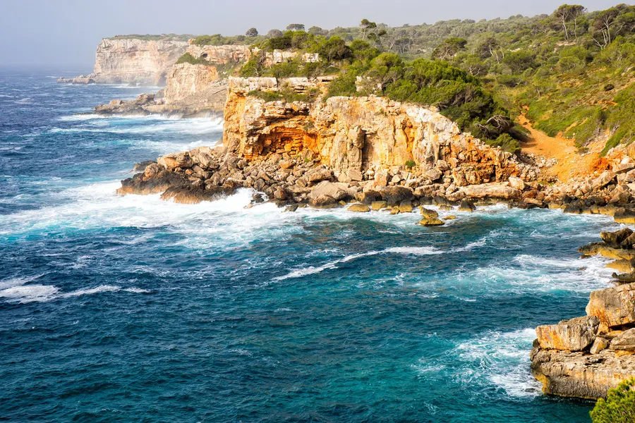 Die zerklüftete Felsküste bei der Cala s'Almunia mit aufgewühltem türkisblauem Wasser und Schaumkronen.
