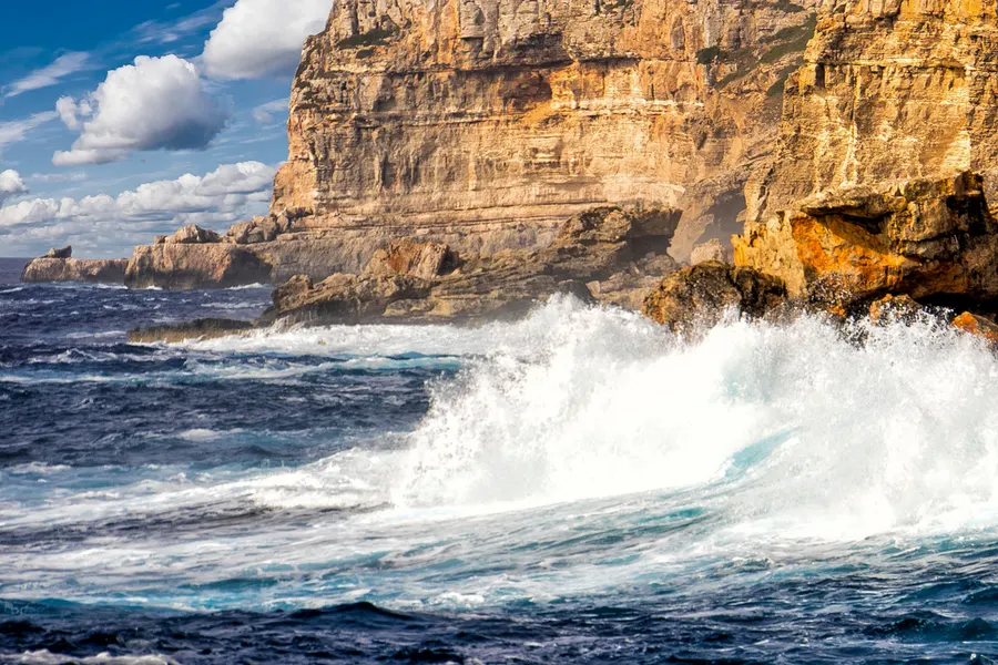 Meterhoch aufspritzende Gischt an einer hellen Felswand der Küste bei Cala s'Almunia.
