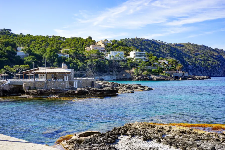 Ein flaches Gebäude mit Terrasse auf einem Felsen im Wasser vor Camp de Mar, dahinter die Bucht und die bewaldeten Hänge mit weißen Häusern.