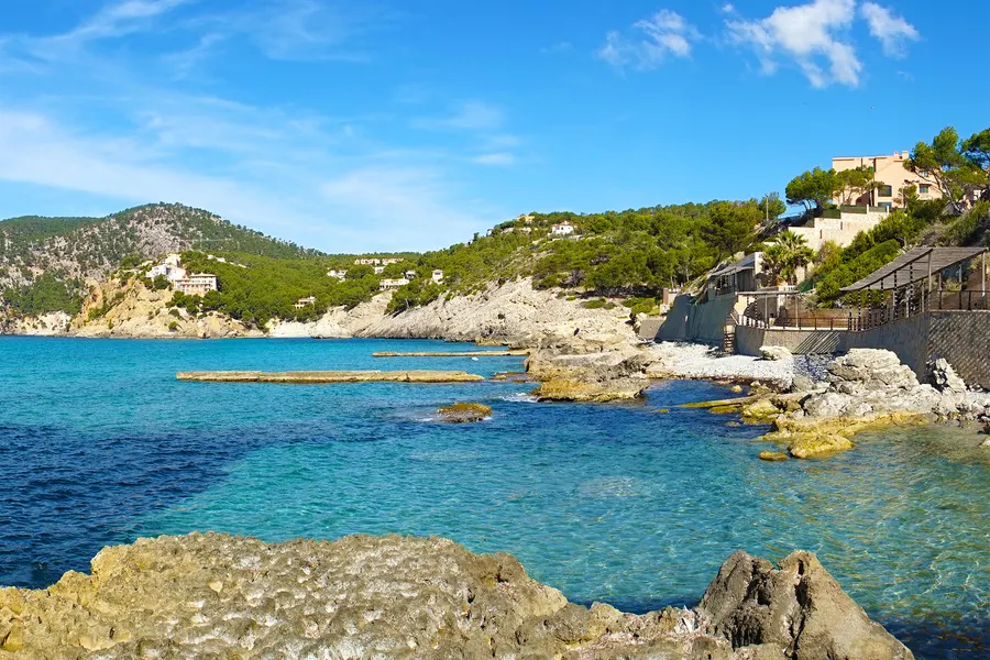 Felsiges Ufer bei Camp de Mar mit klarem türkisem Wasser, einem schmalen Holzsteg entlang der Küste und Häusern zwischen Kiefern am Hang.