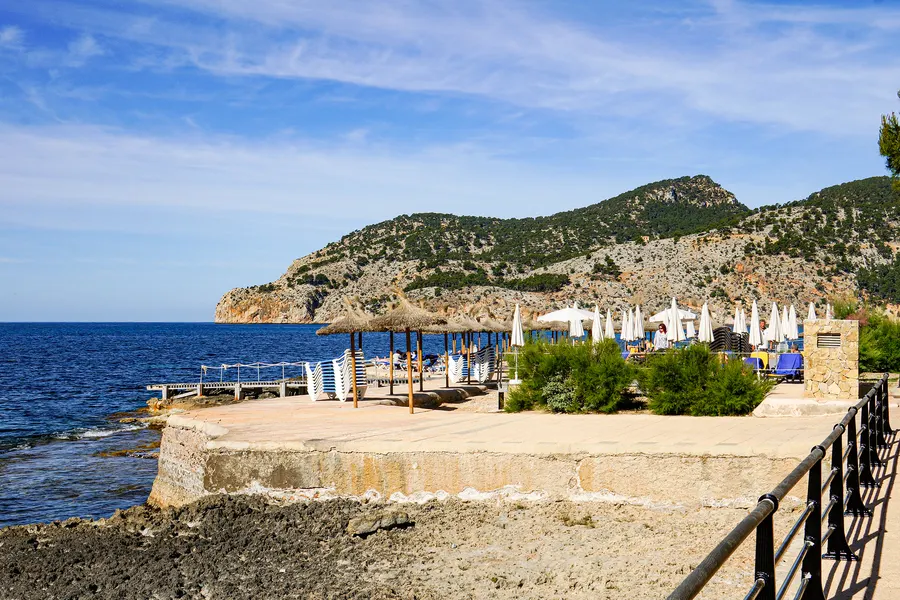 Gemauerte Terrasse mit geschlossenen Sonnenschirmen und Strohdach direkt am Wasser, dahinter das Meer und ein bewaldetes Kap.
