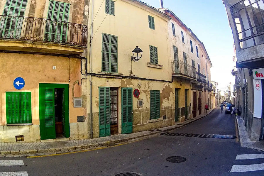 Ruhige Gasse in Campanet mit hellen Häusern, grünen Türen und Fensterläden, verwitterter Fassade und einem Einbahnstraßenschild.
