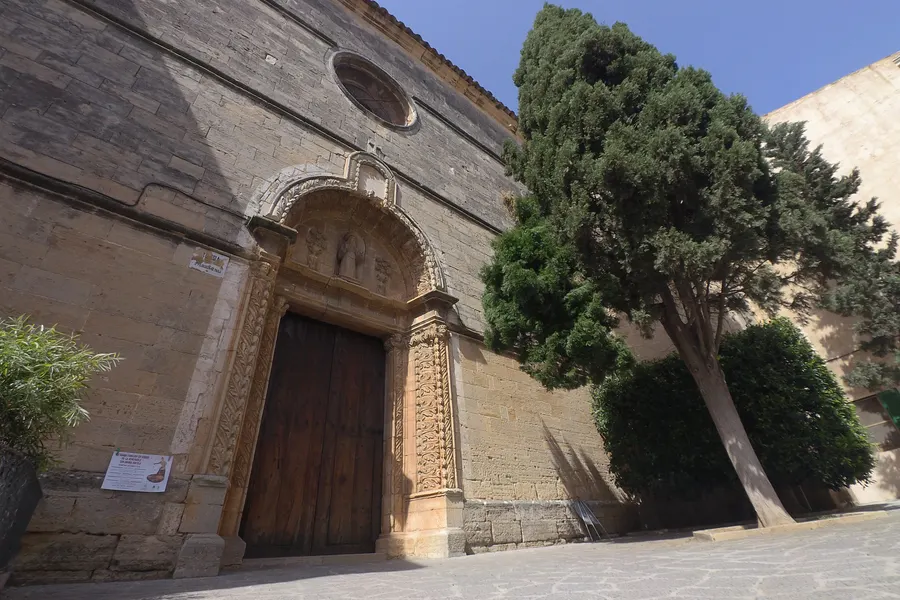 Steinernes Portal der Kirche von Campos mit reich profiliertem Rahmen und schwerer Holztür, daneben eine hohe Zypresse.