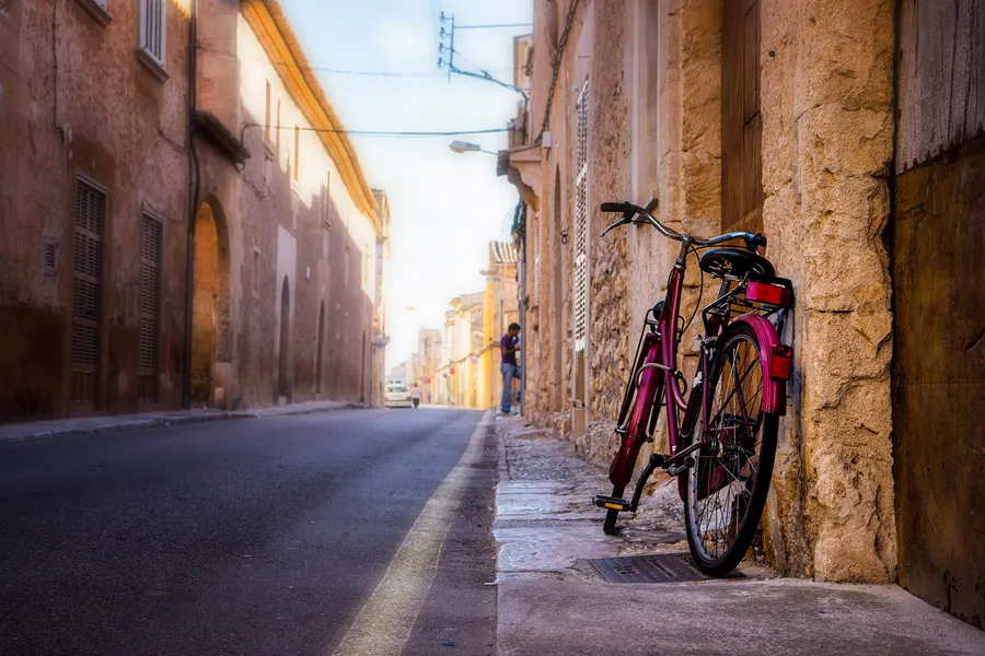 Schmale Gasse in Campos mit einem angelehnten Fahrrad an einer Sandsteinmauer, im Hintergrund eine Person auf der Straße.
