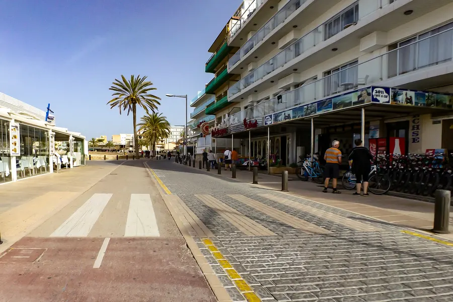 Belebte Geschäftsstraße in Can Pastilla mit Läden, Fahrradverleih, Palmen und Passanten unter blauem Himmel.