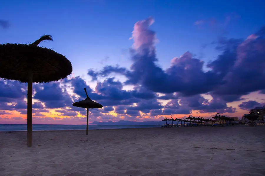 Zwei Strohschirme als dunkle Silhouetten auf leerem Sandstrand vor einem violett und orange gefärbten Abendhimmel, am Horizont die Bergkette der Tramuntana und die Lichterkette der Bucht.