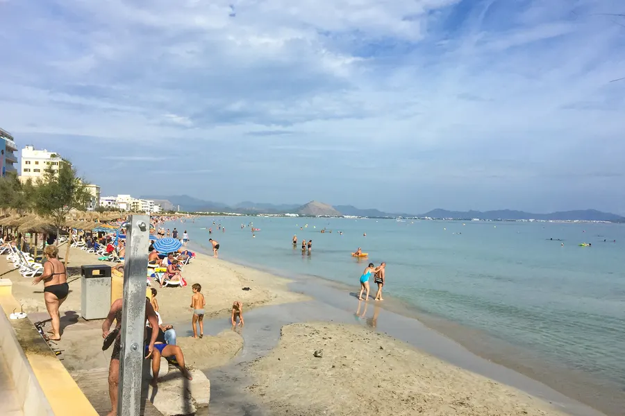 Schmaler Sandstrand im Hochsommer, dicht belegt mit Liegen, Sonnenschirmen und Badenden, links die Hotelzeile mit Strohschirmreihe, rechts flaches türkisfarbenes Wasser mit vielen Schwimmern.
