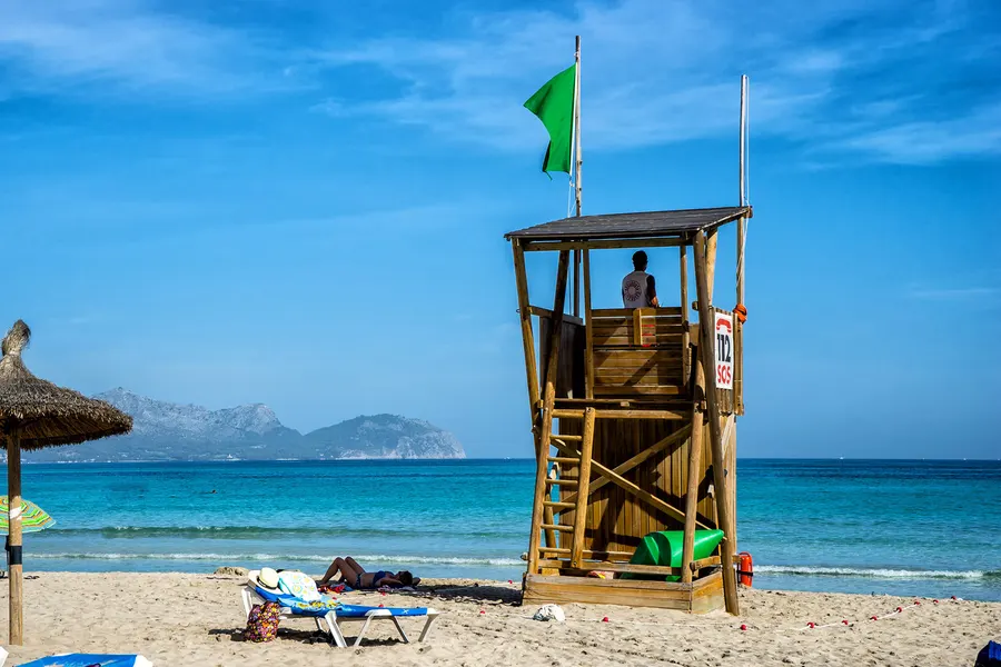 Hölzerner Rettungsschwimmerturm mit grüner Flagge und Notrufschild 112 am Strand, davor eine Liege mit Sonnenhut, dahinter türkisfarbenes Wasser und die Bergsilhouette der Halbinsel Victòria.