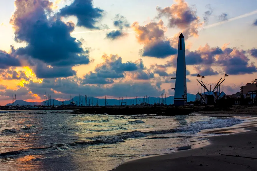 Weißer Obelisk an der Hafeneinfahrt als Silhouette vor einem dramatisch bewölkten Abendhimmel in Orange und Blau, davor die auslaufende Brandung am Sandstrand, im Hintergrund Masten und die Bergkette.
