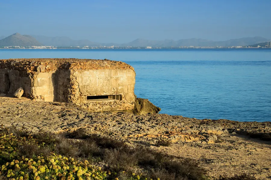 Niedriger Betonbunker mit schmaler Schießscharte auf den Felsplatten direkt am ruhigen, tiefblauen Wasser; am gegenüberliegenden Ufer die Bucht von Alcúdia und dahinter die Berge im Dunst.
